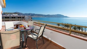 Uma varanda com mesa e cadeiras, com vista para a costa e montanhas ao fundo.