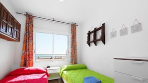 Quarto com duas camas, janela com cortinas, cabeceira de madeira, cómoda branca e decorações nas paredes.