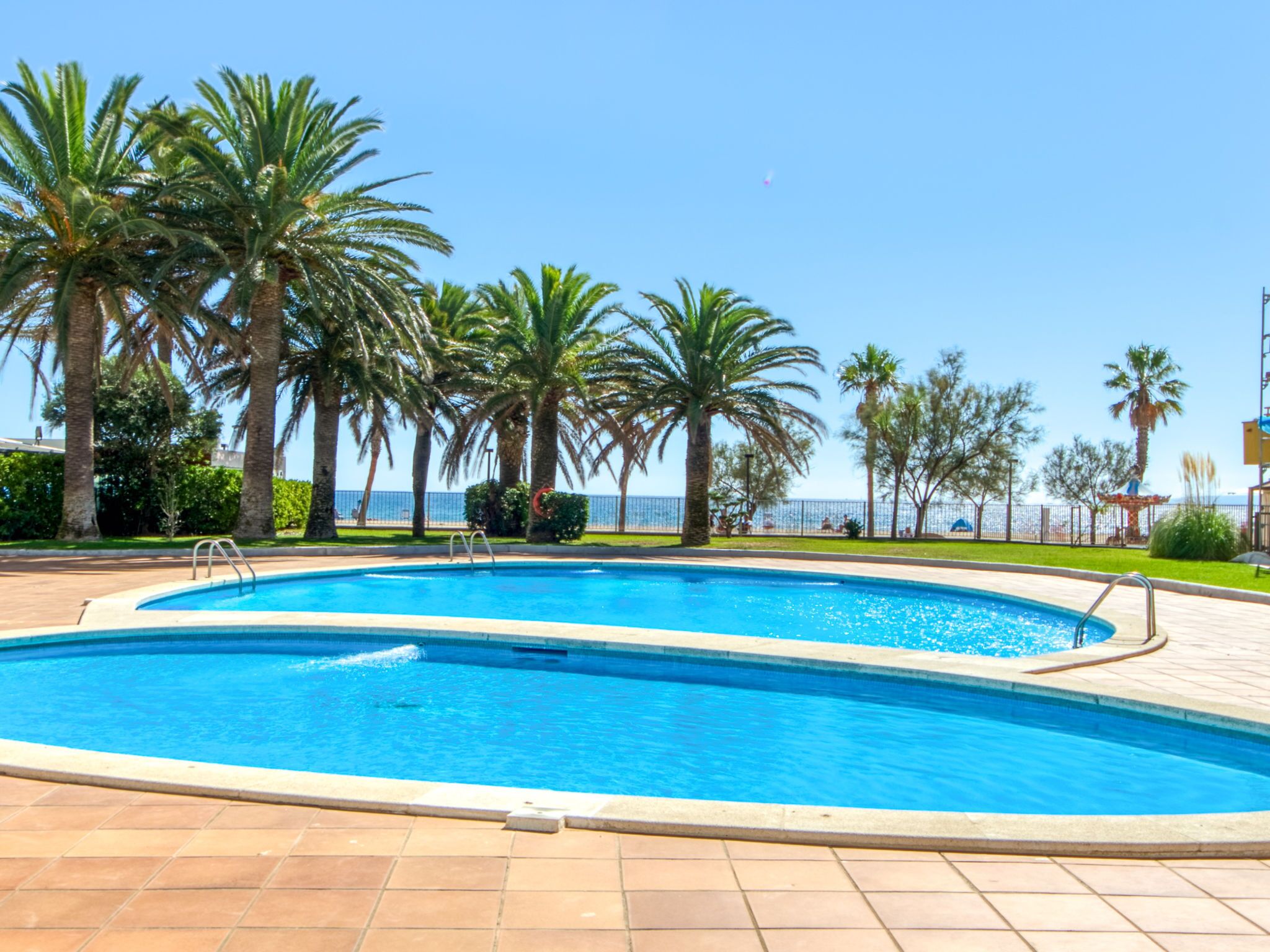 Una piscina con borde de baldosas, rodeada de palmeras y un cielo azul despejado.