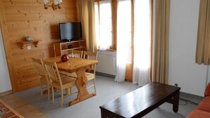 A cozy living room with a wooden dining table, chairs, a dark wooden coffee table, a television on a stand, and a window with curtains.