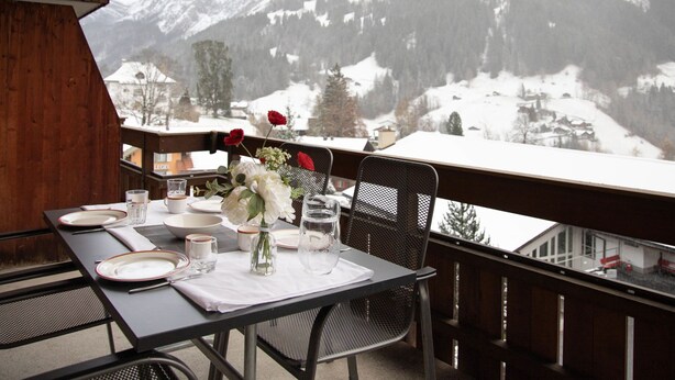 Ein Esstisch ist gedeckt mit Tellern, Gläsern und einer Blumenvase, mit Blick auf eine verschneite Berglandschaft.