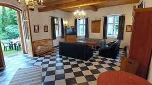 Un salon chaleureux avec un sol à carreaux, des poutres apparentes en bois au plafond, un téléviseur et une vue sur l’extérieur.
