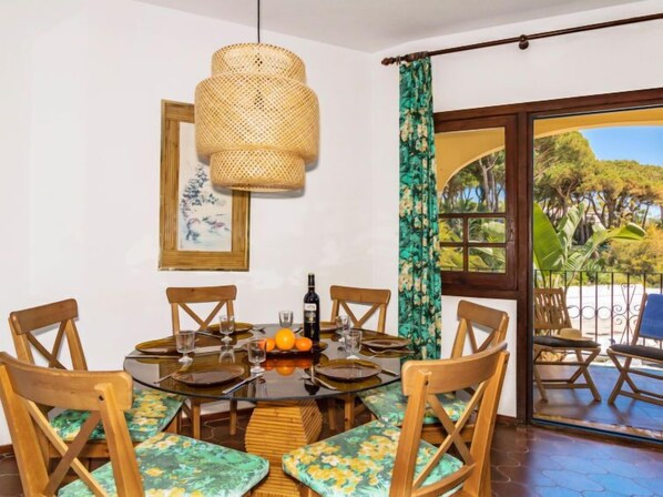 A dining area with a round table, wooden chairs, a woven light fixture, a framed artwork, and a view of the outdoors through a glass door. - Cabopino by Interhome (Marbella)