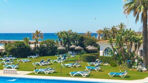 Zona de piscina con sillones reclinables y sombrillas, rodeada de palmeras y vista al océano.