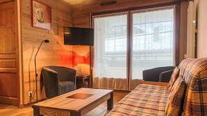 Ein gemütliches Wohnzimmer mit Holzboden, einer Couch, einem Sessel, einem Fernseher und einem Fenster mit Vorhängen.