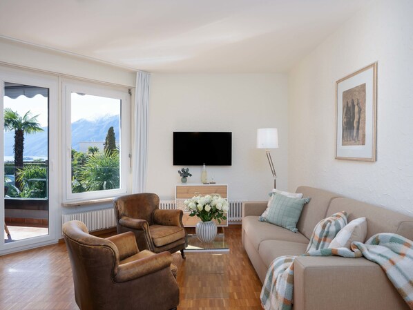 A living room with a sofa, armchair, and a TV. There is a window with a view of mountains and greenery. - Junior Suite Classic - Isole di Brissago by Interhome (Ascona)
