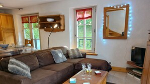 Ein gemütliches Wohnzimmer mit einem braunen Ecksofa, einem Couchtisch aus Holz mit Weingläsern, einem Fernseher und einer Küchenzeile inklusive Holzbodenregal und roten Vorhängen.