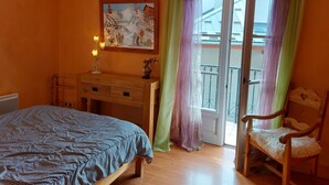 Ein Schlafzimmer mit Bett, Schreibtisch, Stuhl, einem Gemälde und einem Balkon mit Vorhängen.