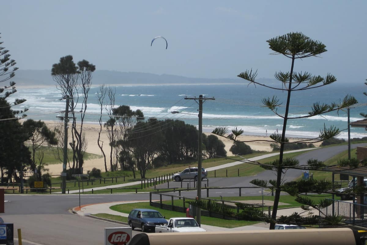 Une route côtière offrant une vue sur la plage, des voitures garées le long du bord de la route et, au loin, un pratiquant de kitesurf.