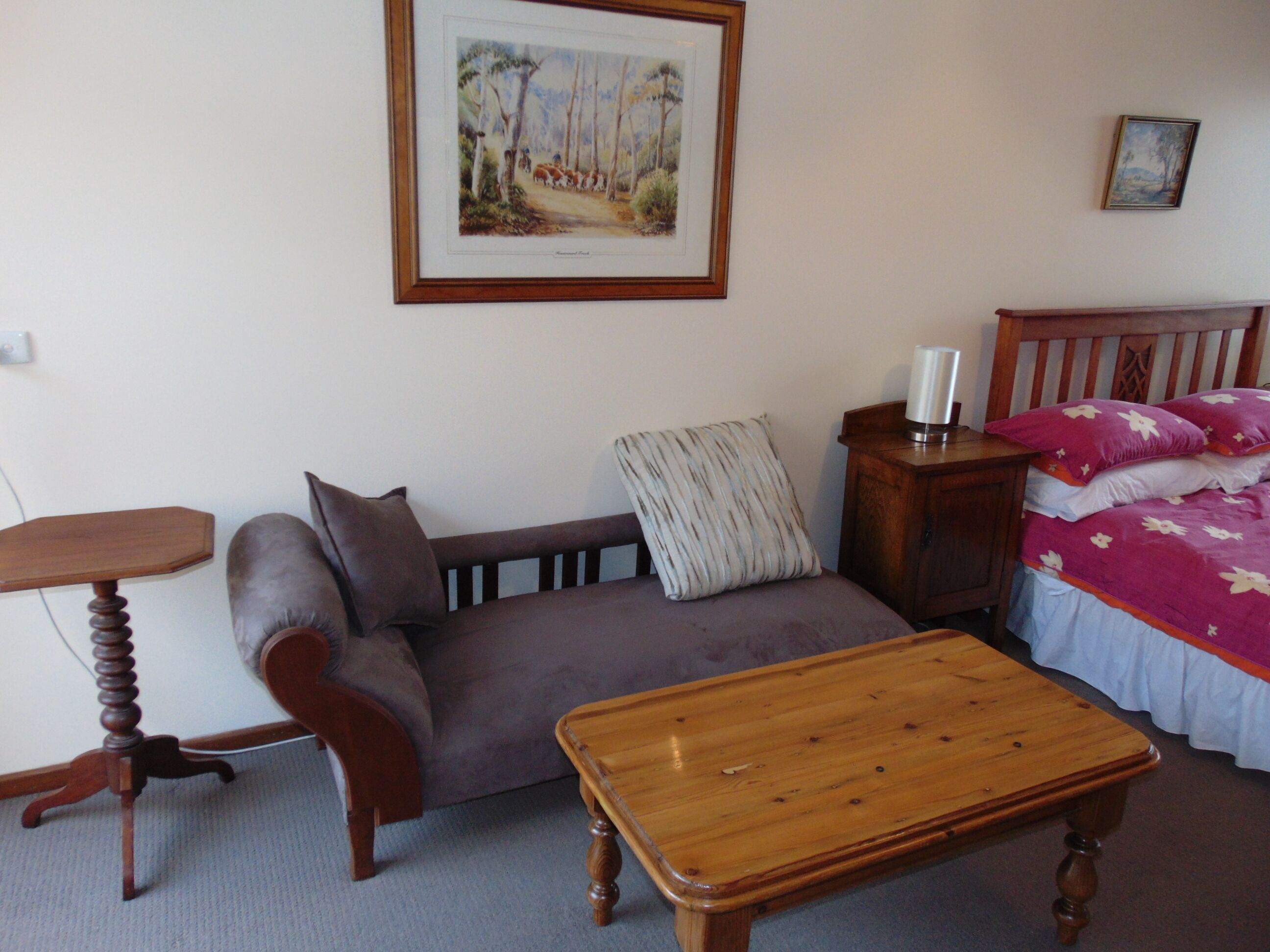 A room with a wooden table, a sofa, a bed with a pink floral bedspread, and a framed picture on the wall.