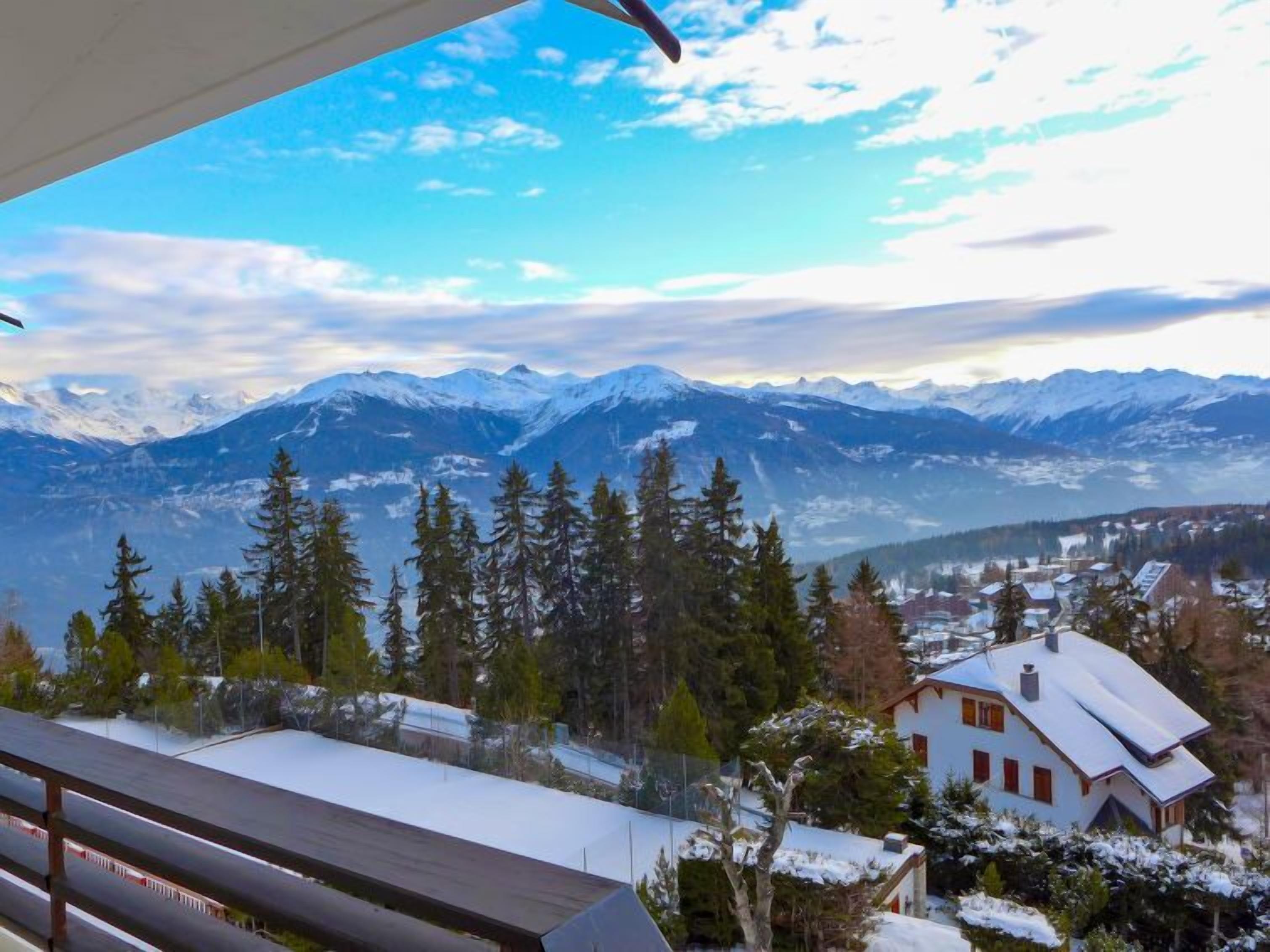 Une vue sur une montagne aux sommets enneigés, une ville située dans la vallée et une maison dont le toit est recouvert de neige.