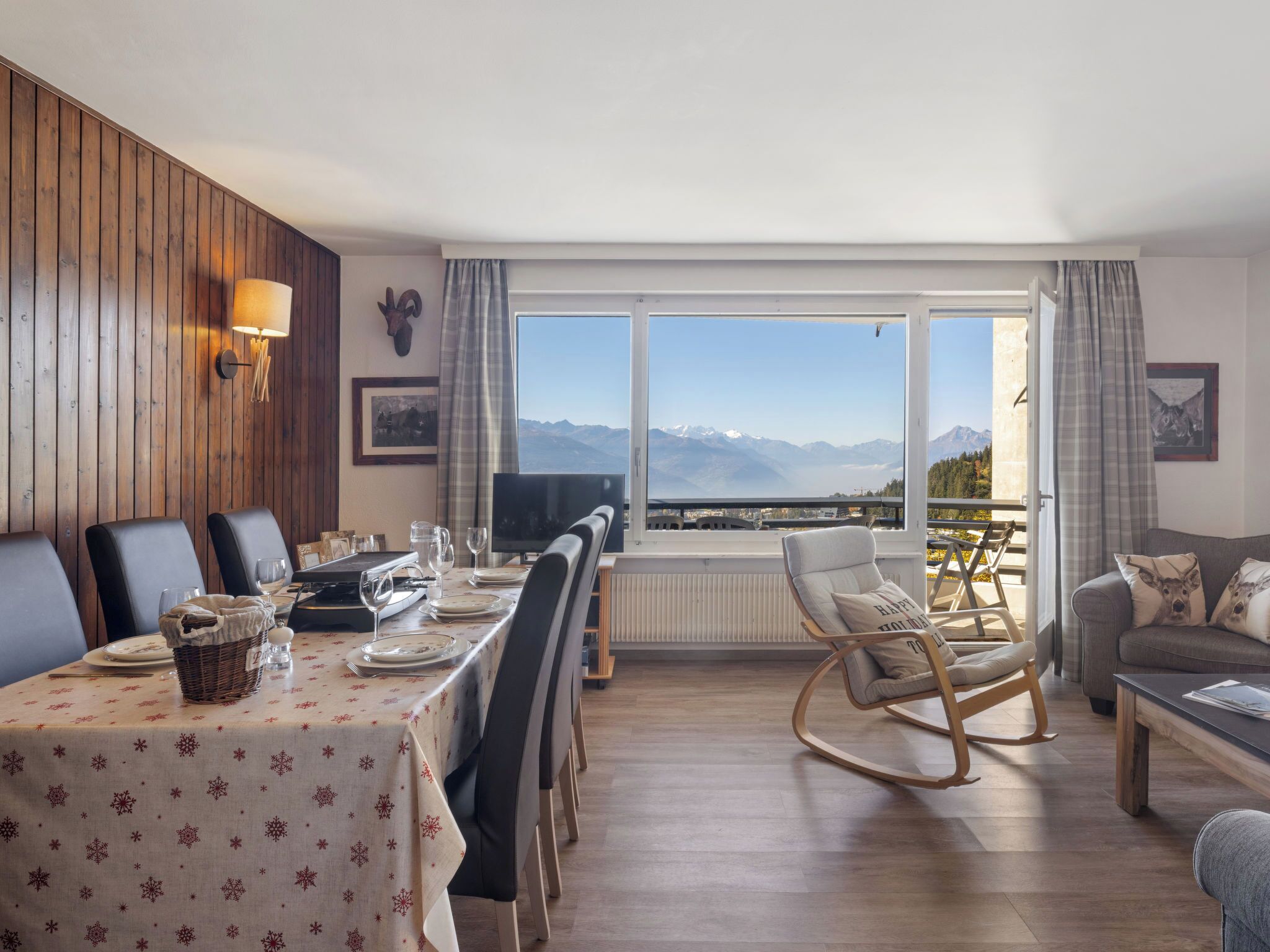 Un coin repas avec une table dressée pour quatre personnes, un téléviseur et une vue sur les montagnes grâce à de grandes fenêtres.