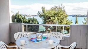 Une table ronde entourée de chaises blanches, sur un balcon donnant sur un lac et des arbres.