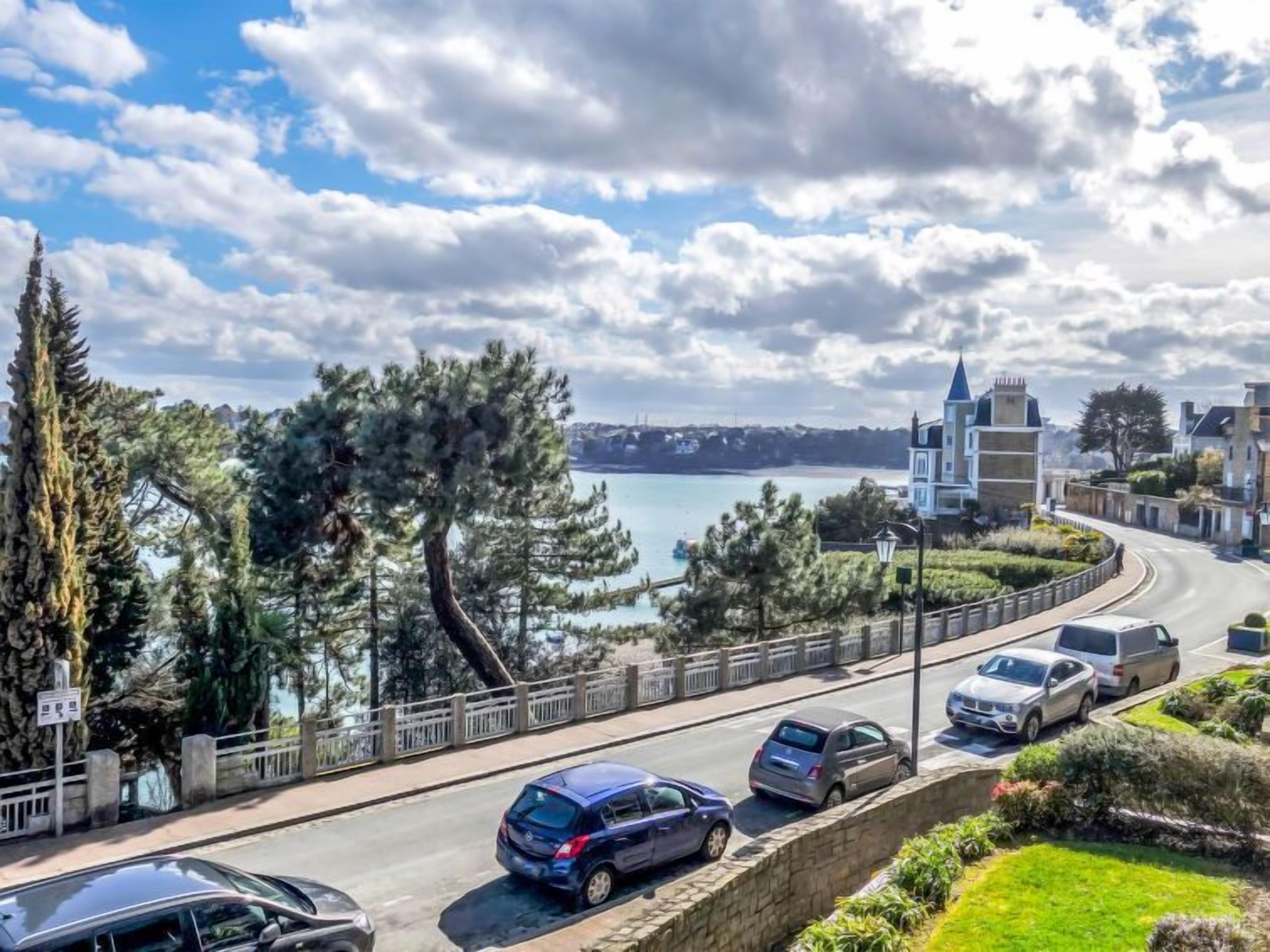 A coastal road with parked cars, a stone wall, and a view of the sea and buildings in the distance.