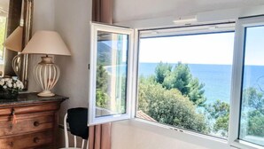 Ein Zimmer mit einem Holzkasten, einer Lampe, einem Stuhl und einem Fenster mit Blick auf Bäume und das Meer.