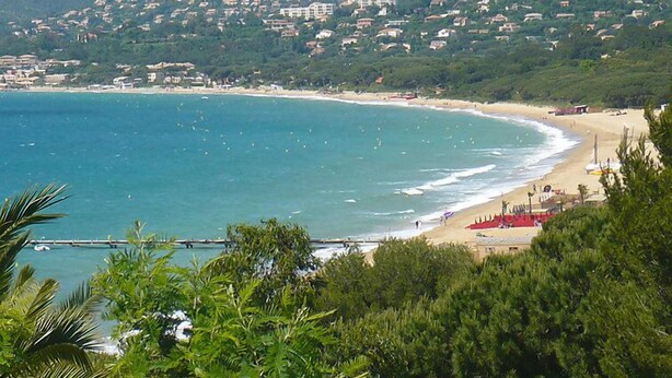 Eine Küstenansicht mit Strand, einer Mole und einer Siedlung am Hang.