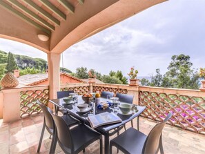 Un espace extérieur abrité, aménagé pour les repas, avec une table dressée pour quatre personnes, entourée de chaises, et offrant une vue panoramique sur un paysage verdoyant avec des arbres et des collines au loin.