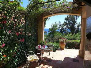 Une terrasse extérieure avec une table et deux chaises, entourée d’une végétation luxuriante et offrant une vue sur la mer.