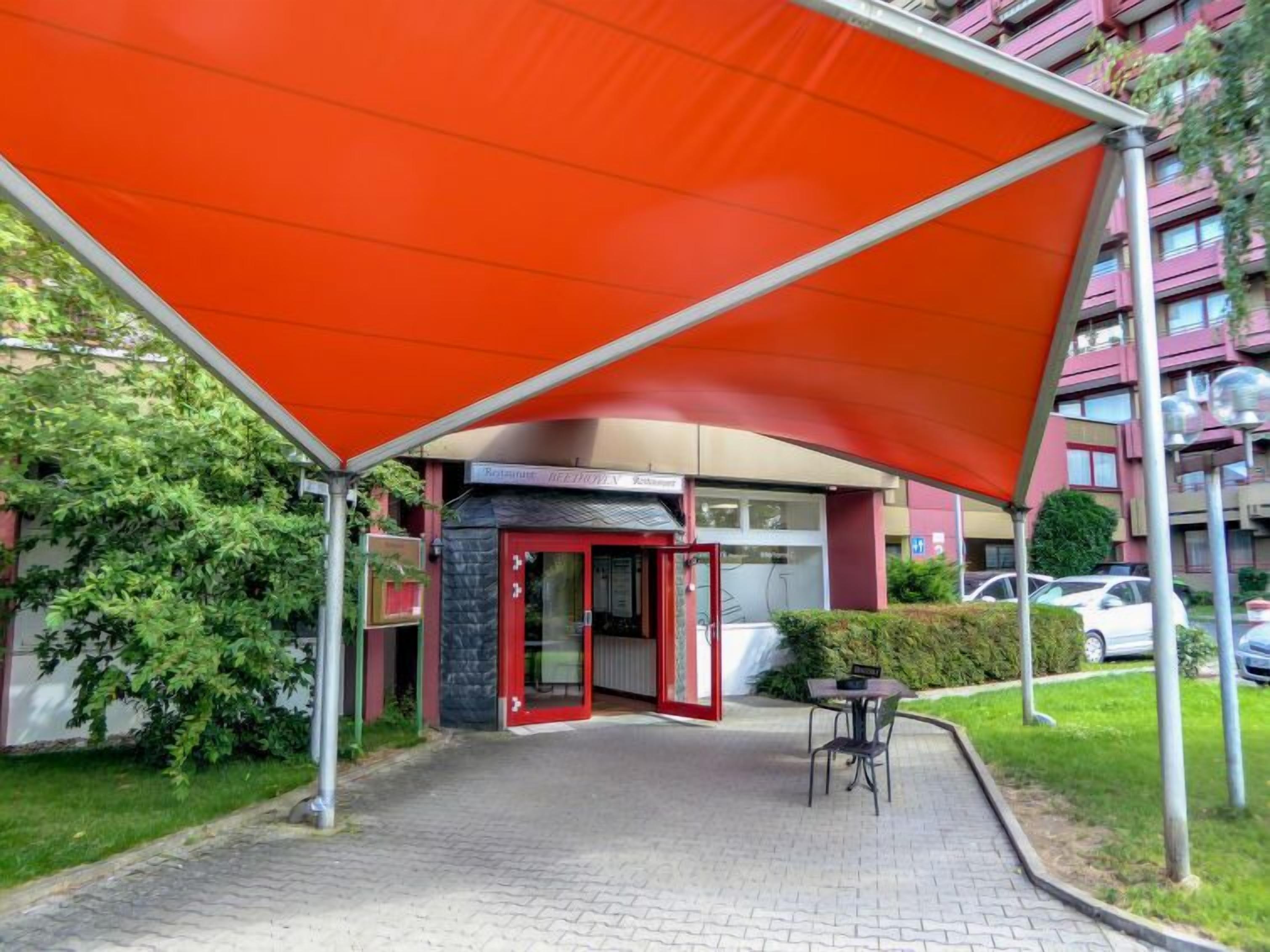 Un espace extérieur abrité, doté d’un auvent rouge, d’une entrée avec porte vitrée et d’une petite table entourée de chaises.