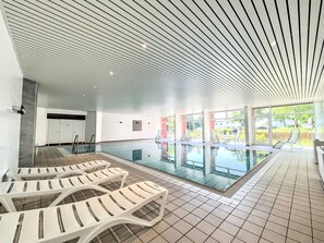 Ein moderner, überdachter Pool mit Glaswänden, weißen Liegestühlen und Bodenfliesen.