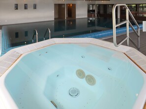 Una amplia piscina interior con superficie de agua cristalina, una escalera metálica y una solana con azulejos alrededor de la piscina.