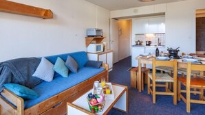 Ein kompakter Wohnbereich mit einem blauen Sofa, einem Holzesstisch und einer Küchenzeile im Hintergrund.
