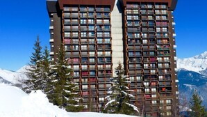 Ein Hochhaus-Hotel mit mehreren Balkonen, umgeben von schneebedeckten Bergen und immergrünen Bäumen.