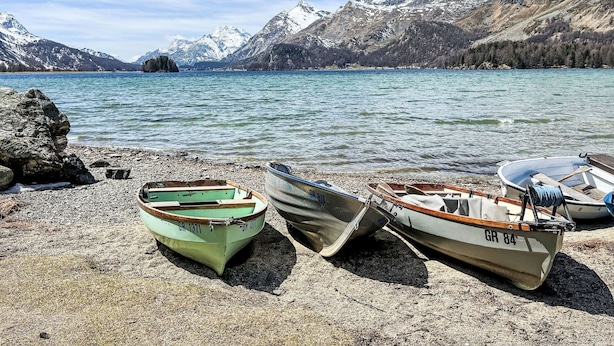 Drei Boote an einer felsigen Uferlinie, neben einem See mit Bergen im Hintergrund.