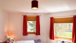Ein Schlafzimmer mit einem Bett, zwei Fenstern mit roten Vorhängen, einem Nachttisch mit Lampe und Blick ins Grüne.