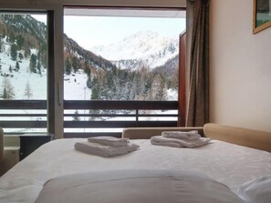 A hotel room with a bed, two folded towels, and a view of snow-covered mountains through a large window.