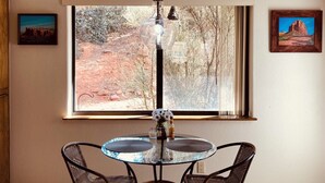 Een eethoek met een ronde glazen tafel, twee rieten stoelen, een hanglamp en uitzicht op een woestijnlandschap door een groot raam.