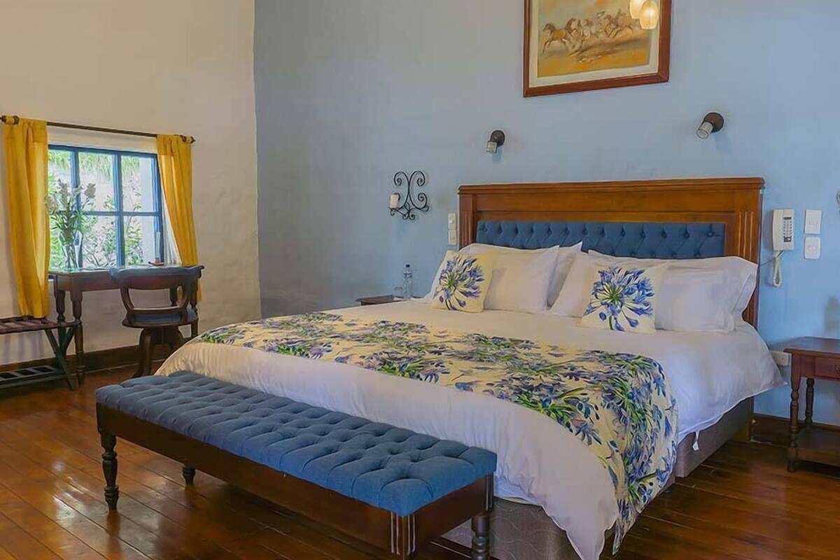 Une chambre à coucher avec un grand lit, un banc en bois, une petite table et une fenêtre avec des rideaux.