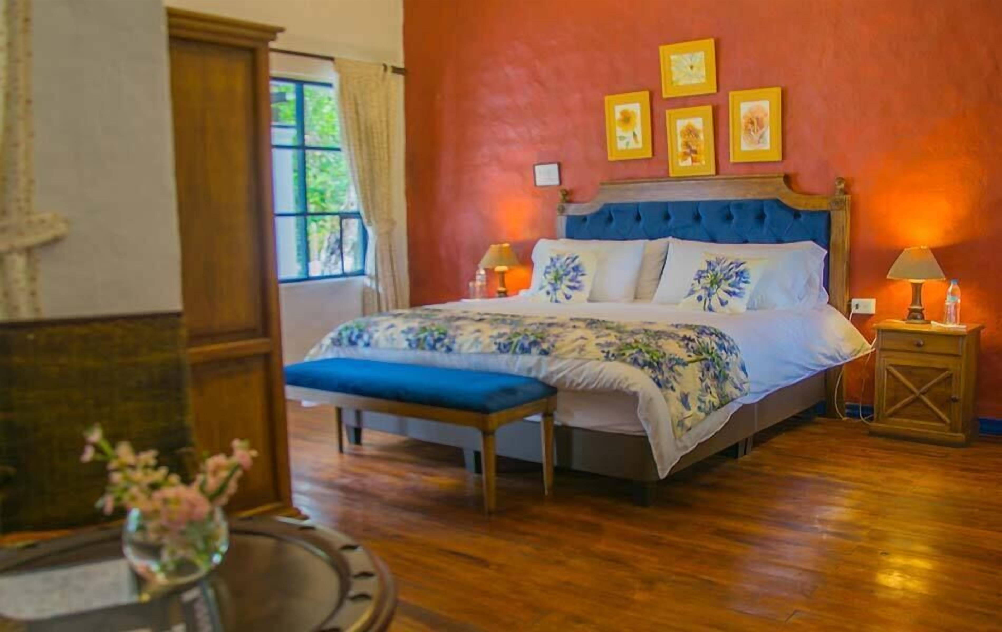 A bedroom with a large bed, a wooden bench, a nightstand with a lamp, and a window with curtains.