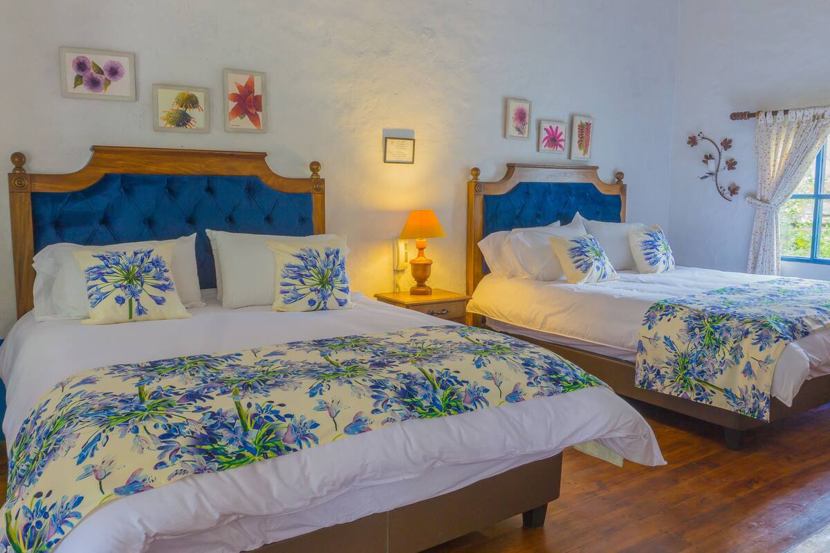 Une chambre d’hôtel avec deux lits, une literie à motifs floraux, une tête de lit en bois, une lampe de chevet et des reproductions botaniques encadrées accrochées au mur.