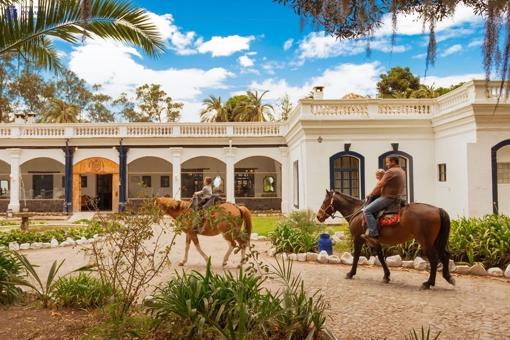 A courtyard with two horses and riders, surrounded by a white building with arches and a balcony.