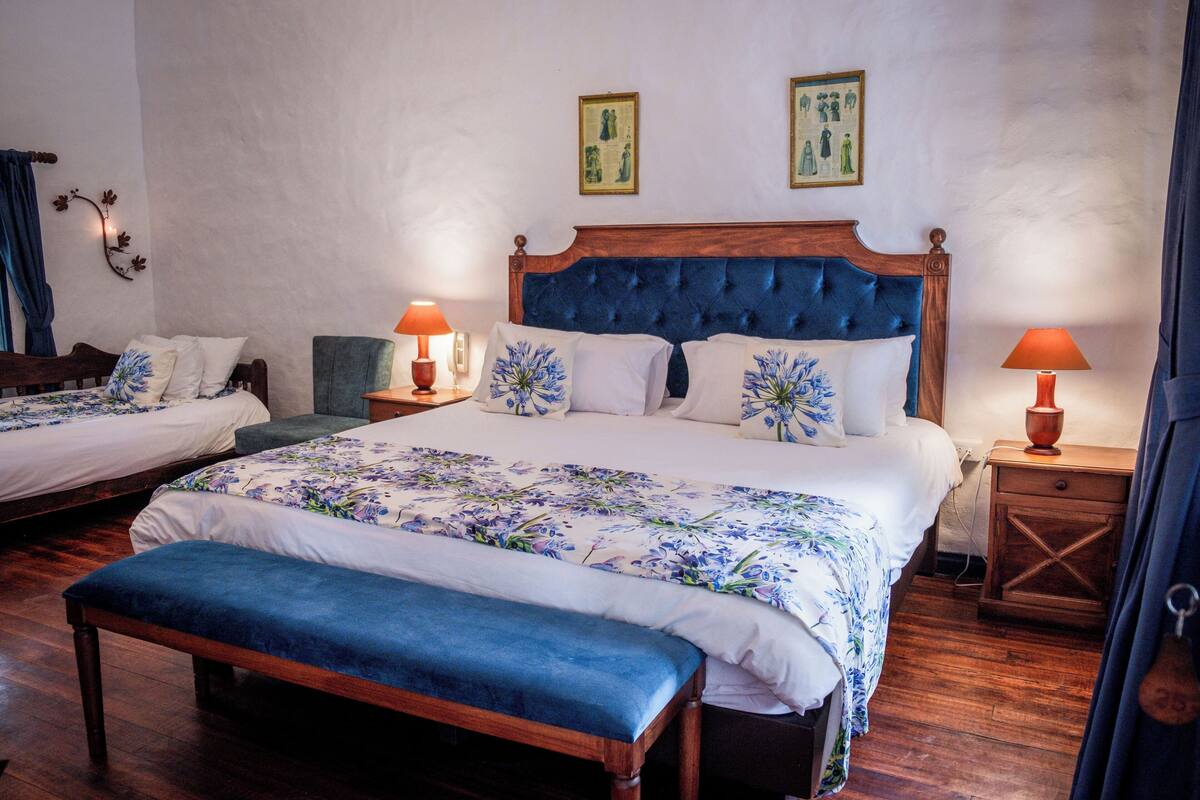 Une chambre double avec deux lits, chacun agrémenté d’une couvre-lit à motifs floraux et de coussins assortis. On y trouve des tables de chevet avec des lampes, un banc et une table de nuit en bois.