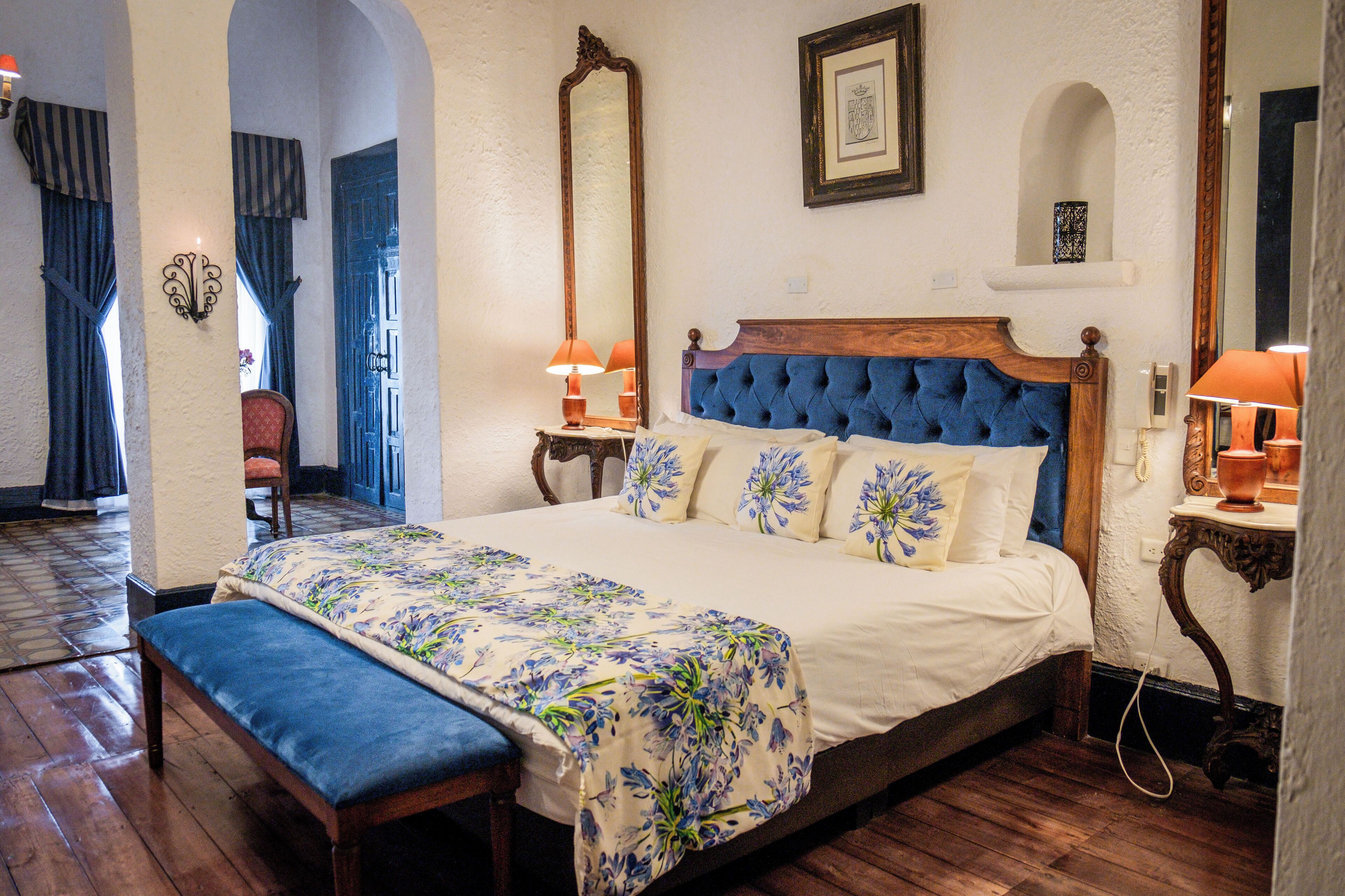 A bedroom with a large bed, wooden headboard, and floral bedding. There are two bedside tables with lamps, a bench, and a chair.