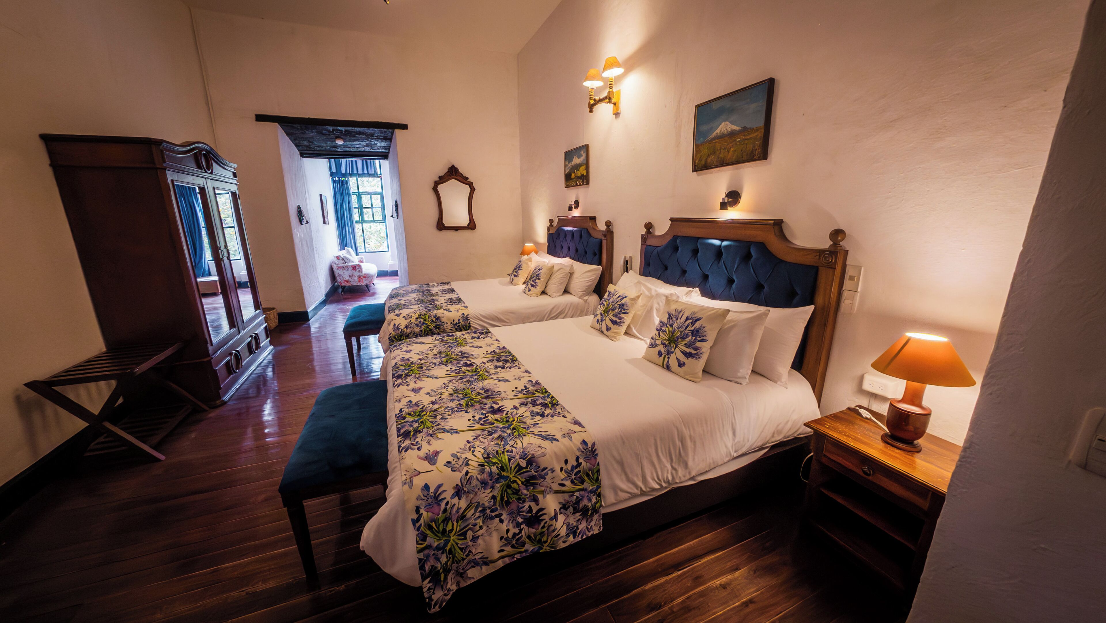 A hotel room with two beds, a wooden floor, a window with curtains, a lamp, and a painting on the wall.