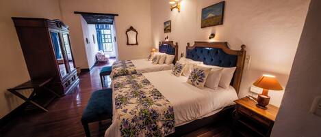 A hotel room with two beds, a wooden floor, a window with curtains, a lamp, and a painting on the wall.