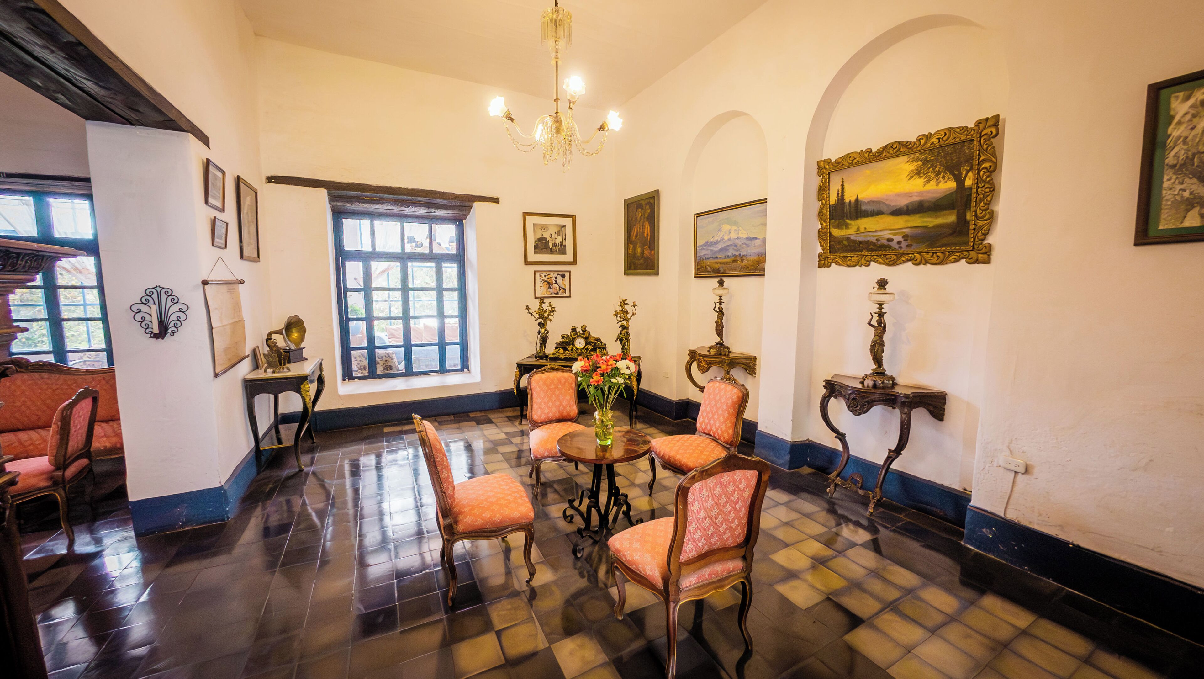 Una habitación con suelo a cuadros, una araña, una mesa con un jarrón de flores y varias sillas.