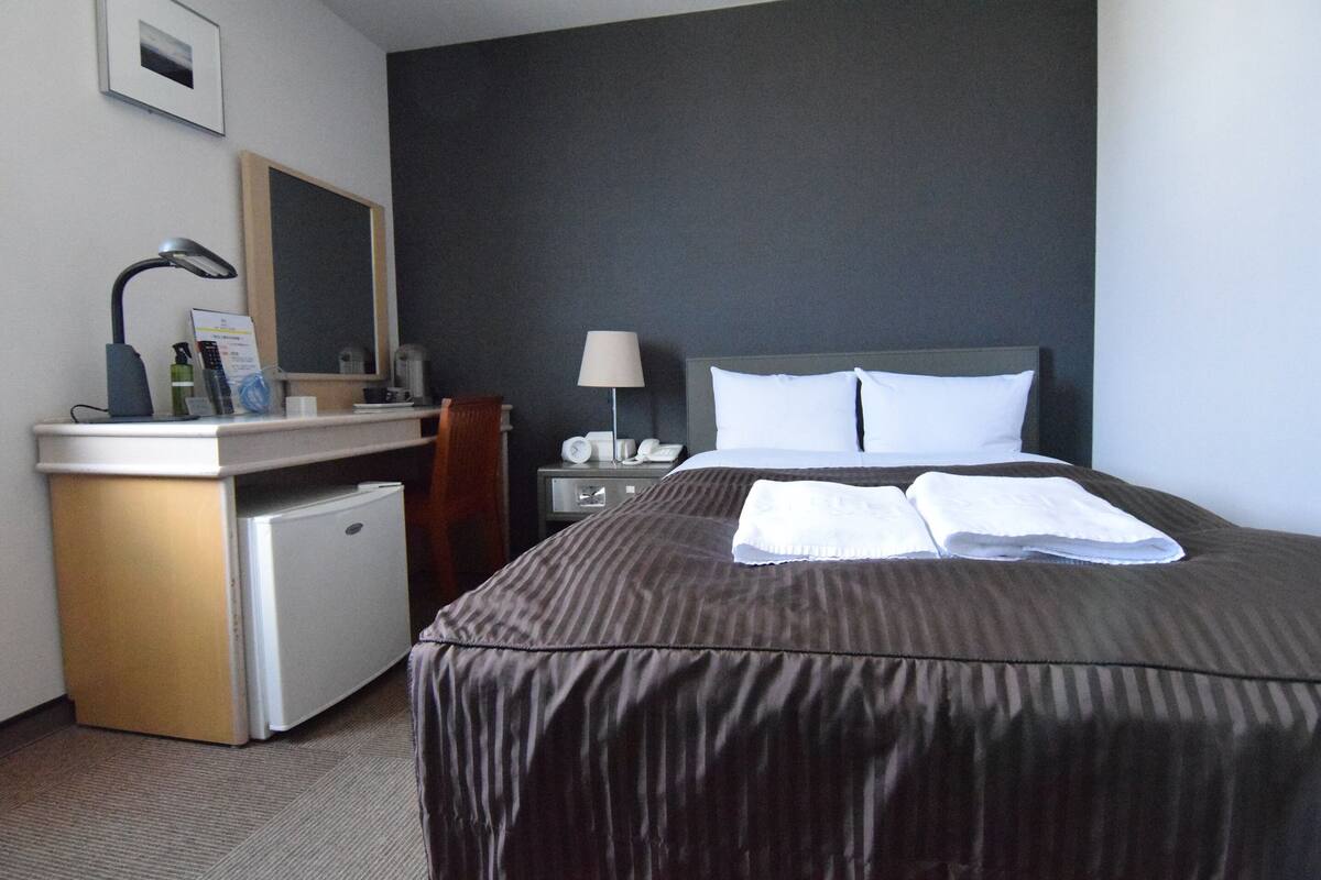 A hotel room with a bed, a desk with a lamp, a small refrigerator, a nightstand with a lamp, and a mirror on the wall.