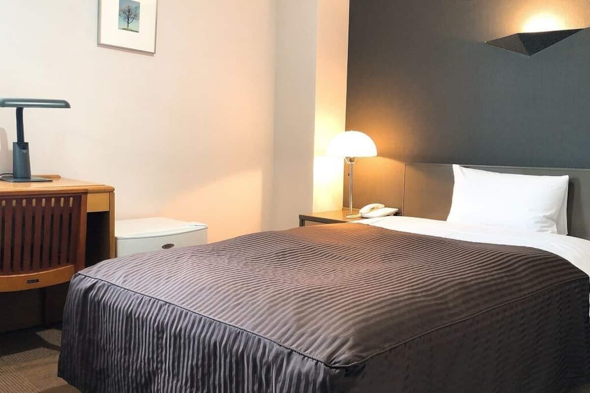 A hotel room with a bed, bedside table, lamp, and a desk with a chair.