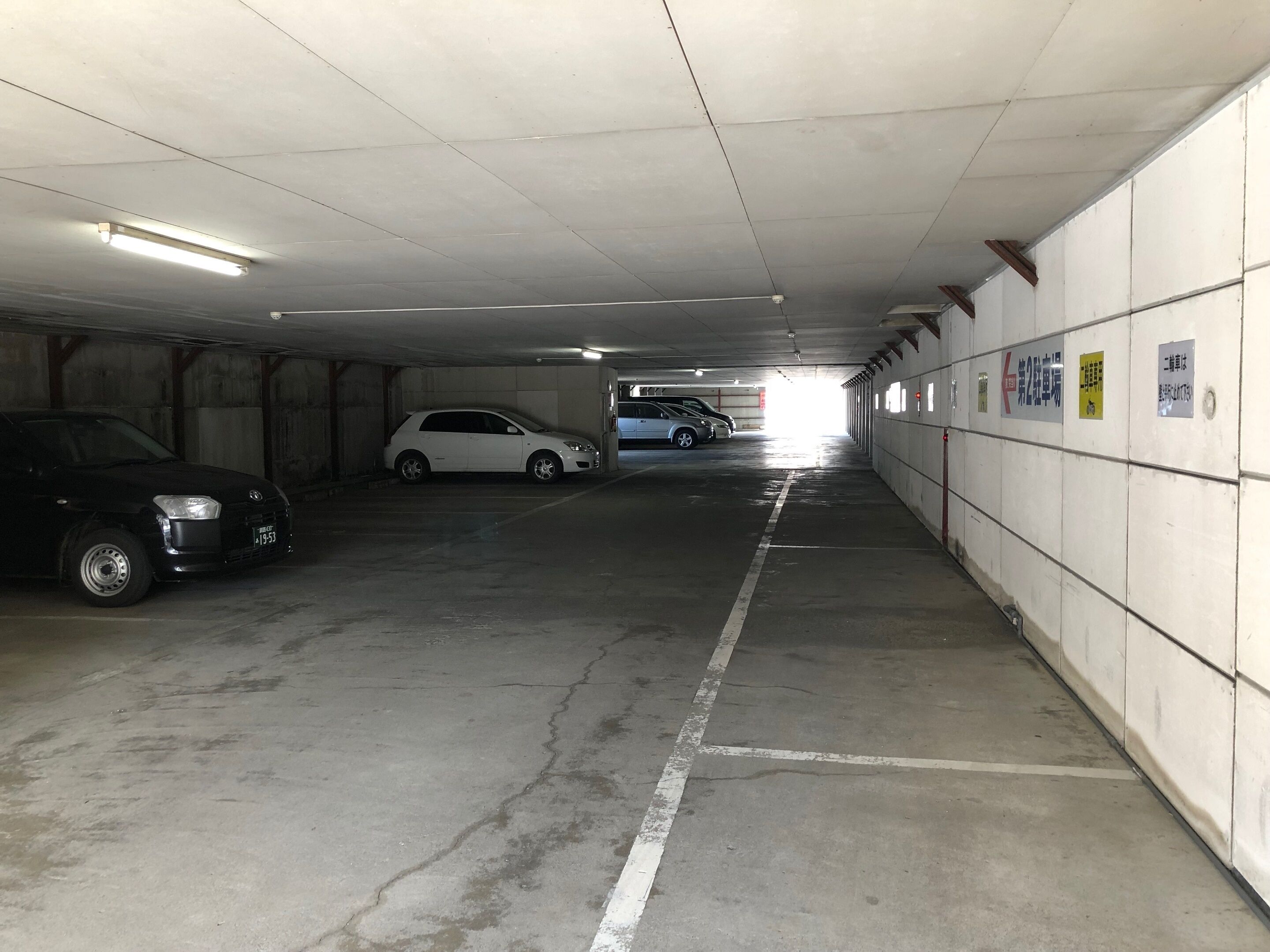 Un aparcamiento con suelos y paredes de hormigón, iluminación fluorescente y varios coches estacionados.
