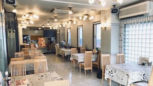 A restaurant with multiple tables and chairs, ceiling lights, and a kitchen area in the background. - Hotel New White House (Rumoi)