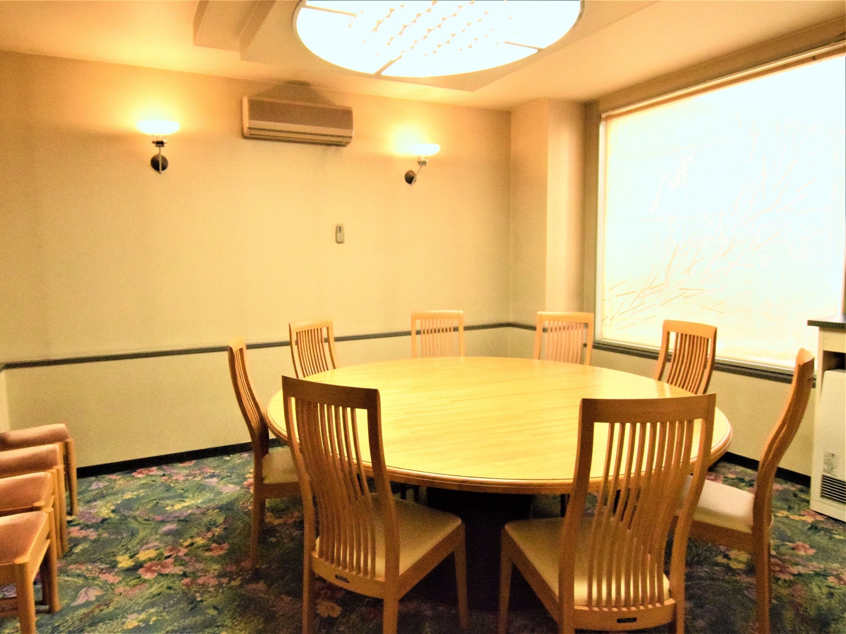 A conference room with a round table, six wooden chairs, two wall-mounted lamps, an air conditioning unit, and a large window with a view.