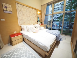 Una camera da letto moderna con un letto grande, un comodino, un tavolino rosso, un tappeto decorato e una finestra con vista su edifici e alberi.