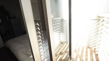 A room with a bed, a sliding glass door leading to a balcony with a brick floor and white railing.