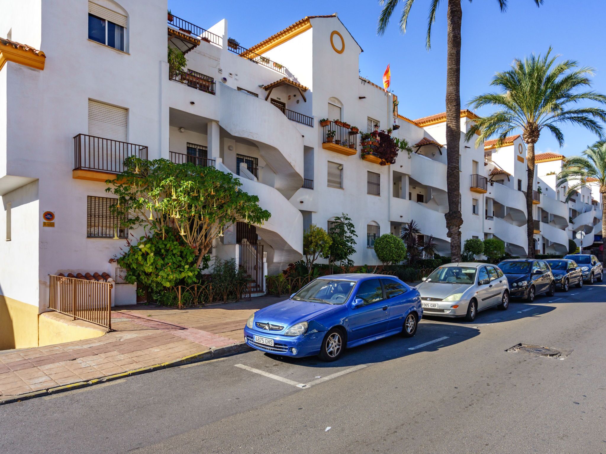 A row of white buildings with balconies, parked cars, and palm trees.