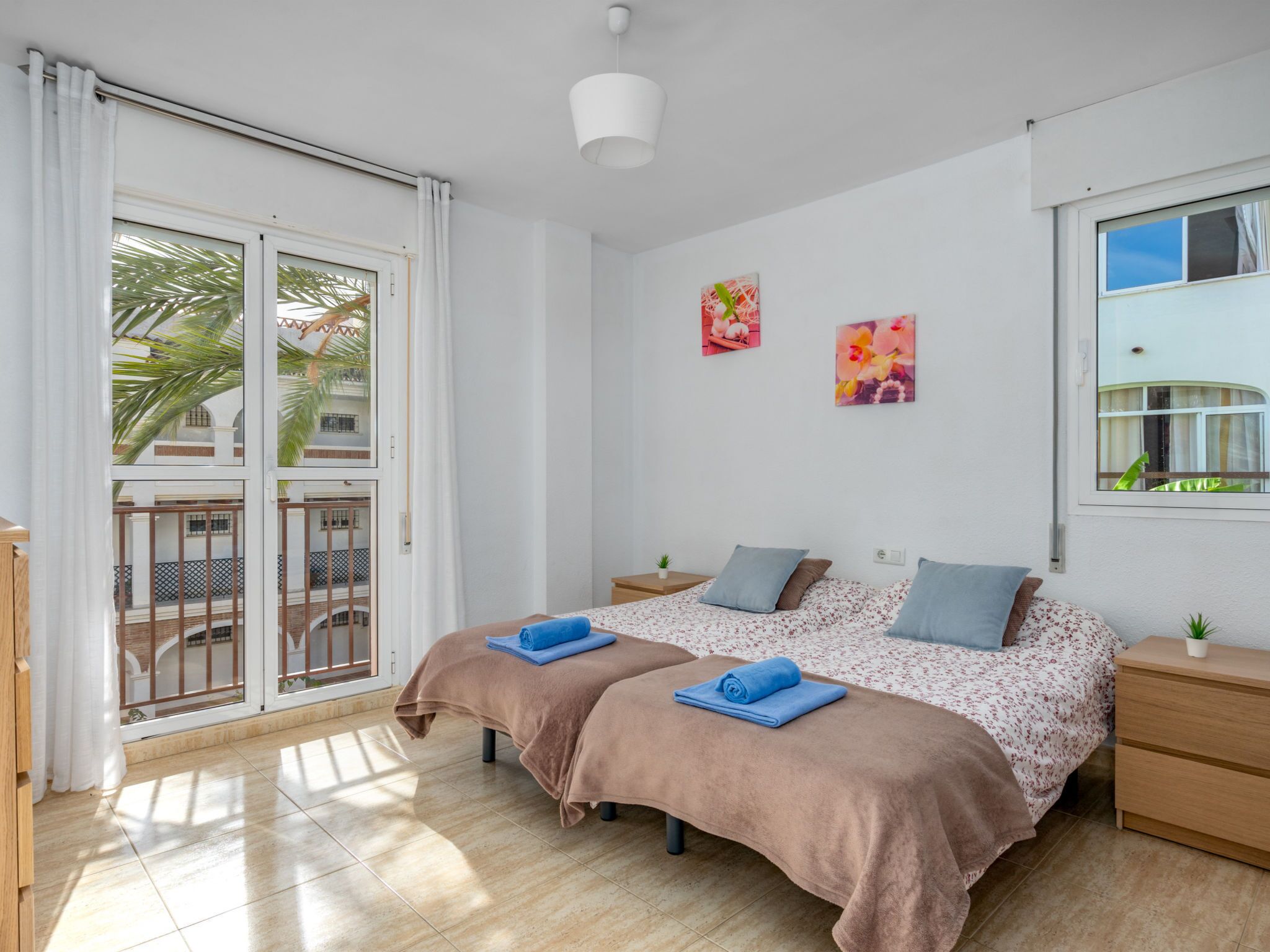 A bedroom with two beds, a balcony, and a view of palm trees.