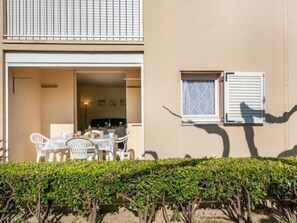 An open patio with a dining table and chairs, a small window with shutters, and a hedge in the foreground.